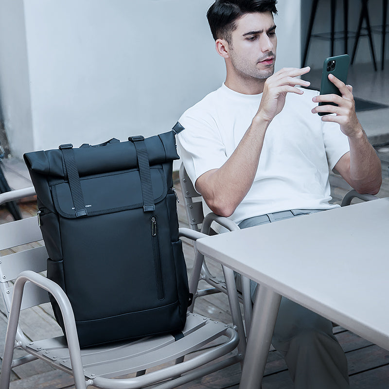 Switch black large capacity daily business travel multifunctional backpack with roll-top closure and front zipper pocket, positioned on a chair beside a person using a smartphone.