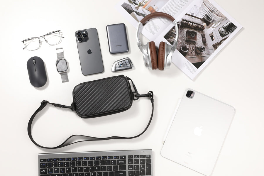 Black anti-theft crossbody bag with diagonal stripe pattern, displayed among travel essentials like smartphone, headphones, and tablet on a clean white surface.