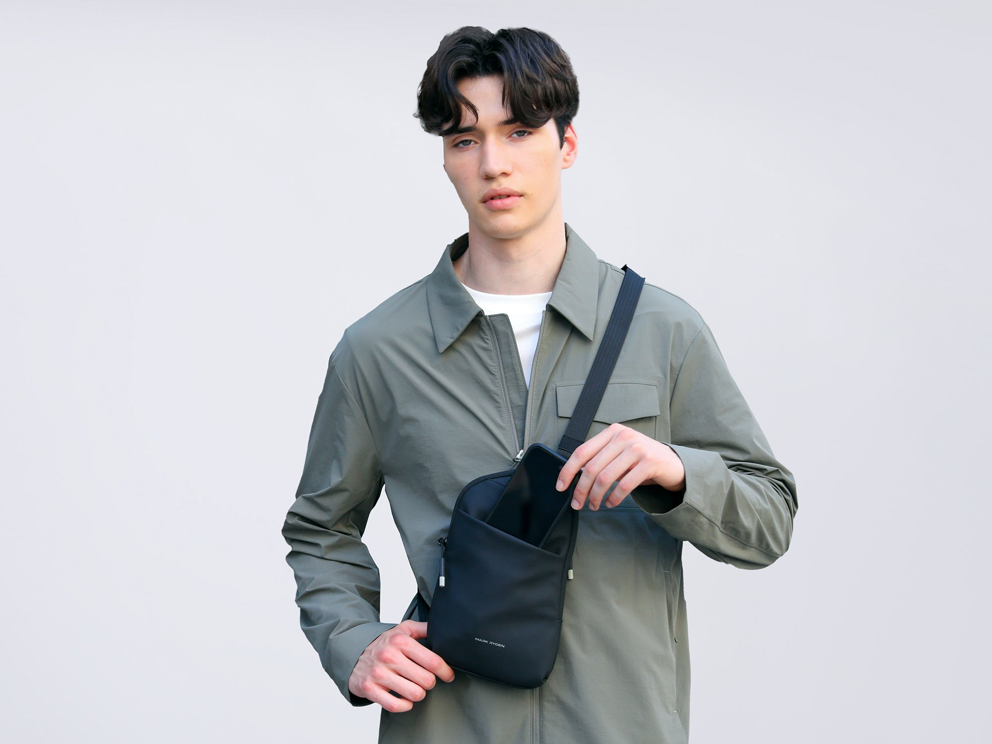 Mark Ryden Global black crossbody bag with adjustable strap, person in green jacket placing smartphone inside