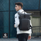 Black large-capacity camera backpack with roll-top closure, worn by a man in a white jacket and black pants, against a modern glass building background, Mark Ryden Global G Series