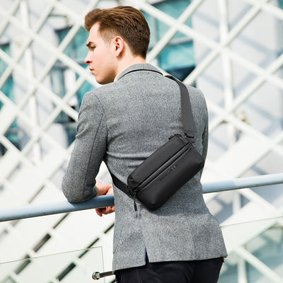 Man wearing black waterproof crossbody bag with multiple compartments over gray blazer, modern architectural backdrop | Mark Ryden Global