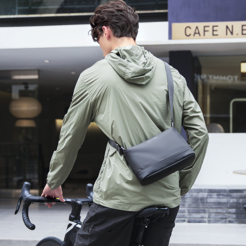 Black lightweight waterproof crossbody bag worn by a man in a green jacket while riding a bike near a café.