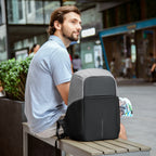 Gray and black Mark Ryden Mocchasio anti-theft backpack with USB charging, resting on a bench in an urban setting.