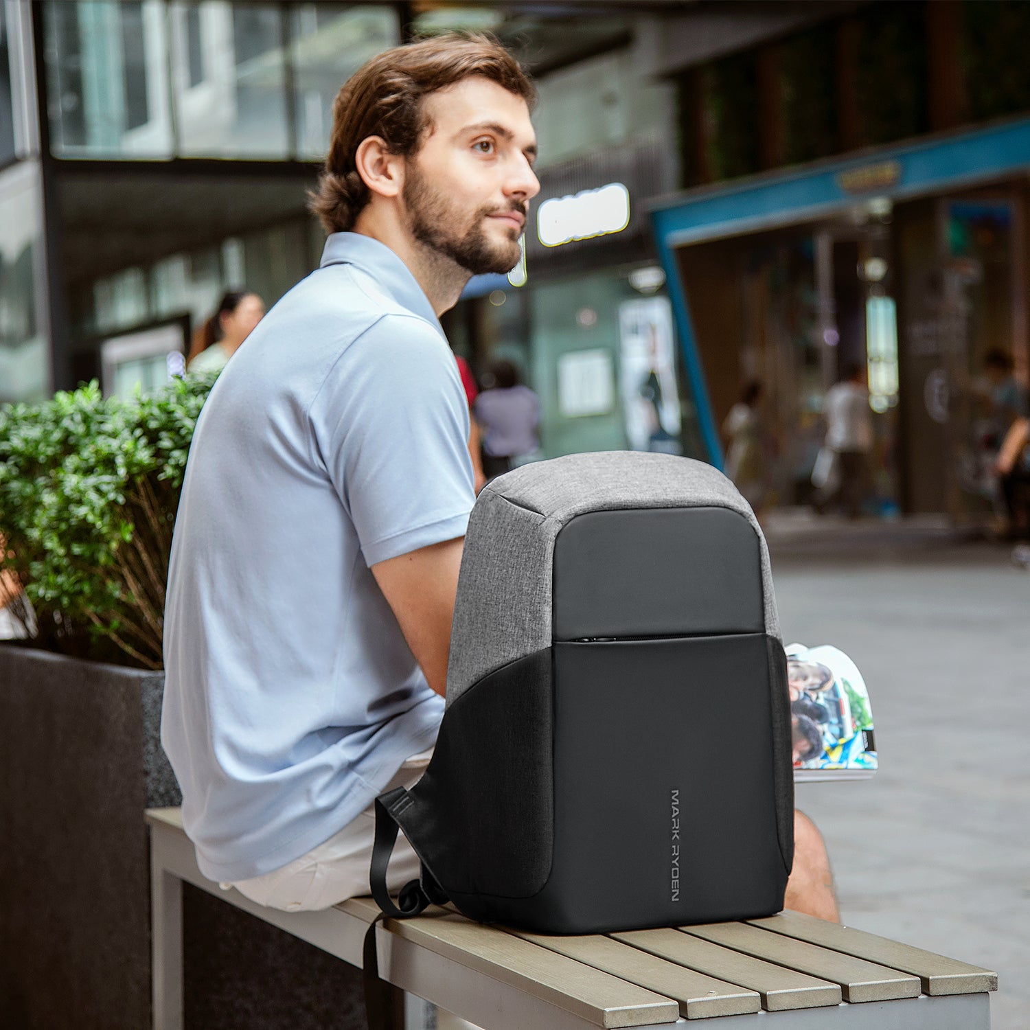 Gray and black Mark Ryden Mocchasio anti-theft backpack with USB charging, resting on a bench in an urban setting.