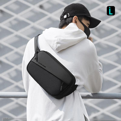 Mark Ryden CrossWear black waterproof crossbody bag with multiple compartments and front zip pocket, worn by person in white hoodie and black cap against grid background.