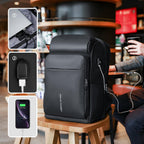 Black Mark Ryden Compacto spacious carry-on laptop backpack with USB charging port, shown with laptop and phone charging in café setting.