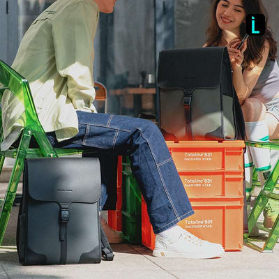 Black MUKE I school style classic business work oxford backpack with flap and buckle, displayed outdoors on orange crates with another backpack in background.
