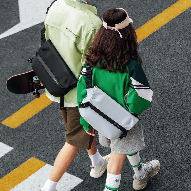 Youthful white and black messenger bags with adjustable straps, carried by two people walking on a street with yellow crosswalk lines.