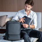 Dark gray ChargeFlex smart backpack with modern sleek design and front zippered pocket, placed beside a man packing a suit jacket on a bed.