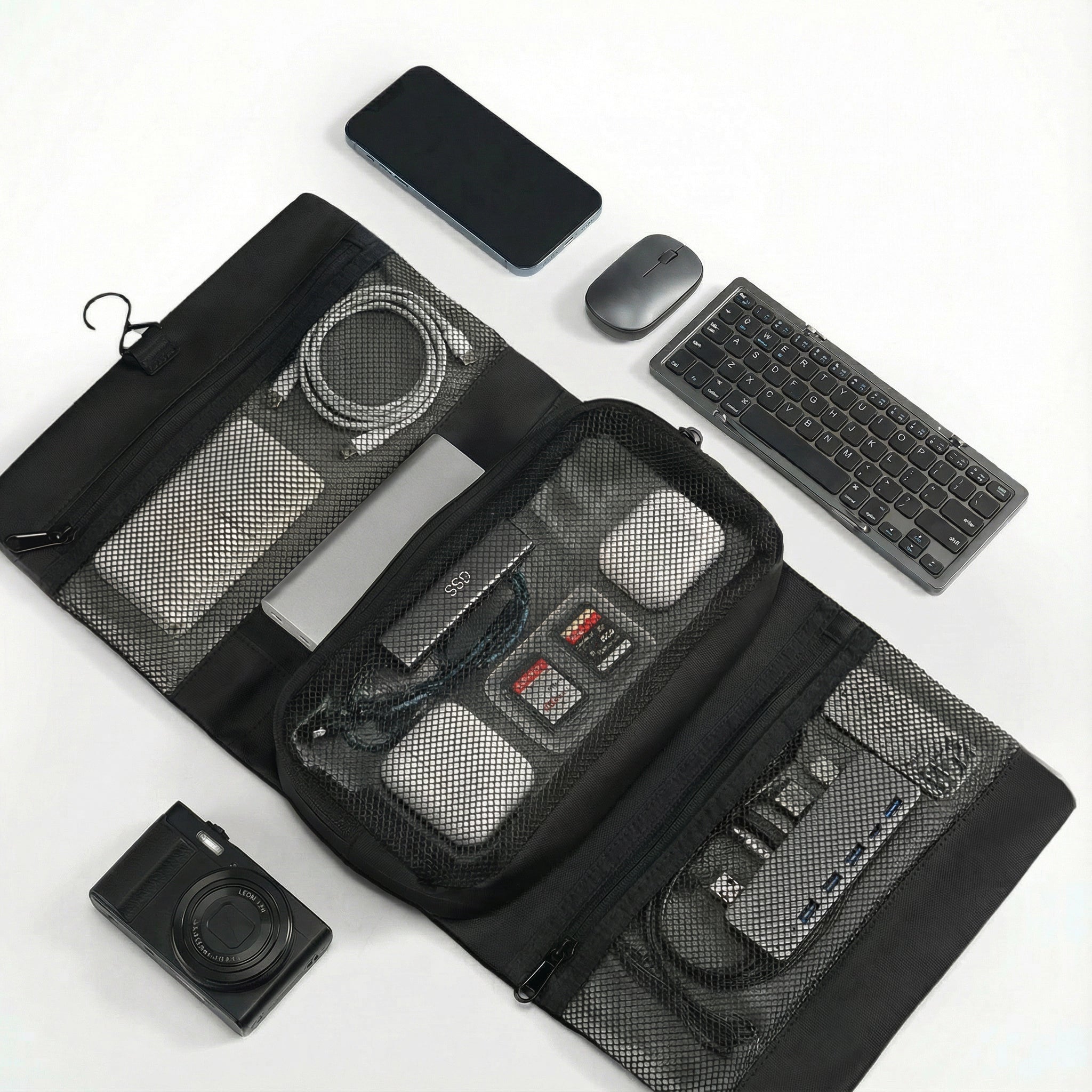 Black MagiClip Wet-Dry Separate Toiletry Bag with mesh compartments holding power bank, cables, SD cards, and small electronics, surrounded by smartphone, mouse, keyboard, and camera.