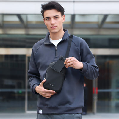 Black Mark Ryden ProCarry business sling bag with front pocket holding smartphone, worn by man in dark blue zip-up sweatshirt against urban backdrop.